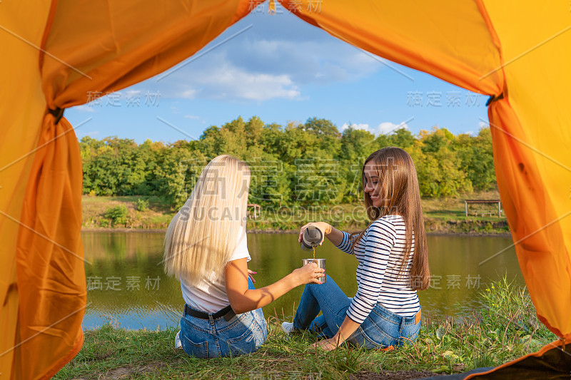 日落时，在湖边露营，从露营帐篷内观看。两个美丽的女孩在帐篷前享受自然，喝着热茶