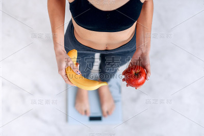 身材完美的运动女性拿着一个黄香蕉和一个红苹果用电子秤测量体重。