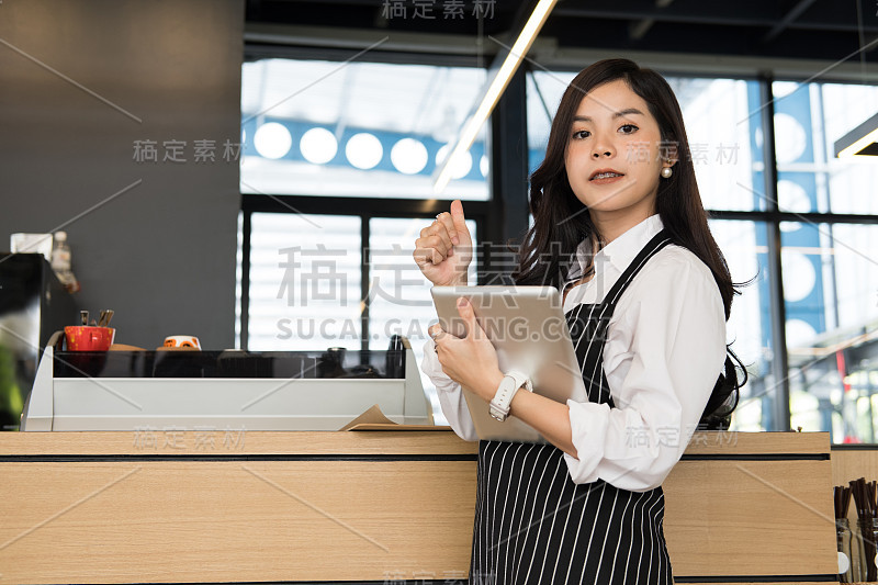 在咖啡店柜台拿着药片的小企业主。女咖啡师穿着围裙在咖啡馆的酒吧使用触控板。餐饮服务、餐厅、创业理念。