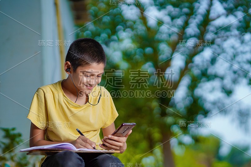 印度一名男孩手持手机，使用手机应用程序进行远程学习，观看在线课程，视频呼叫在应用程序中做笔记，在家学