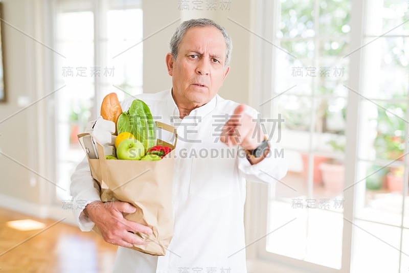 英俊的老男人拿着一纸袋新鲜的食品从超市指向相机和你，手势，积极和自信的手势从前面