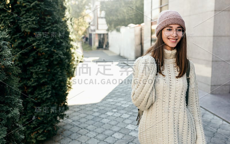 开朗的年轻女子面带微笑，走在城市的大街上，穿着毛衣，背着背包，戴着时髦的透明眼镜。