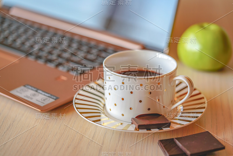 木桌上放着一杯诱人的咖啡，背景是笔记本电脑和苹果。在茶托上放着一块黑巧克力，这是办公室休息时间的近景