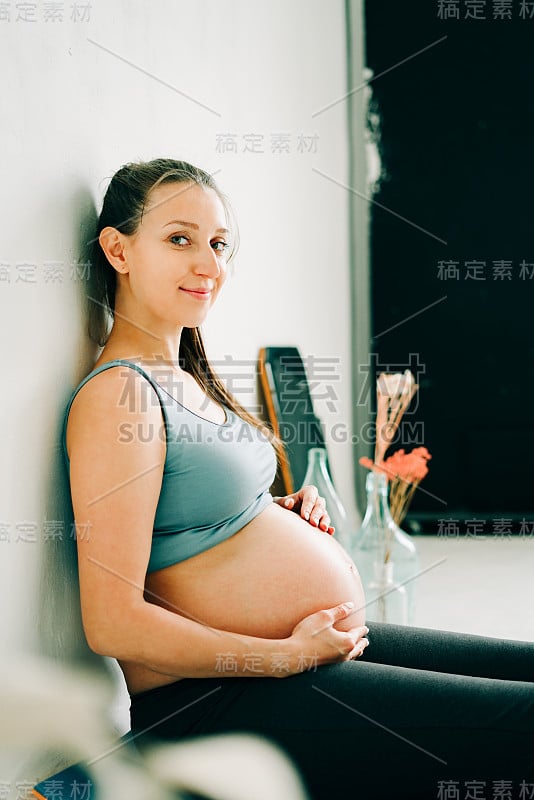 在演播室里，一个年轻漂亮的怀孕女孩穿着运动服坐在地毯上，抱着她的肚子