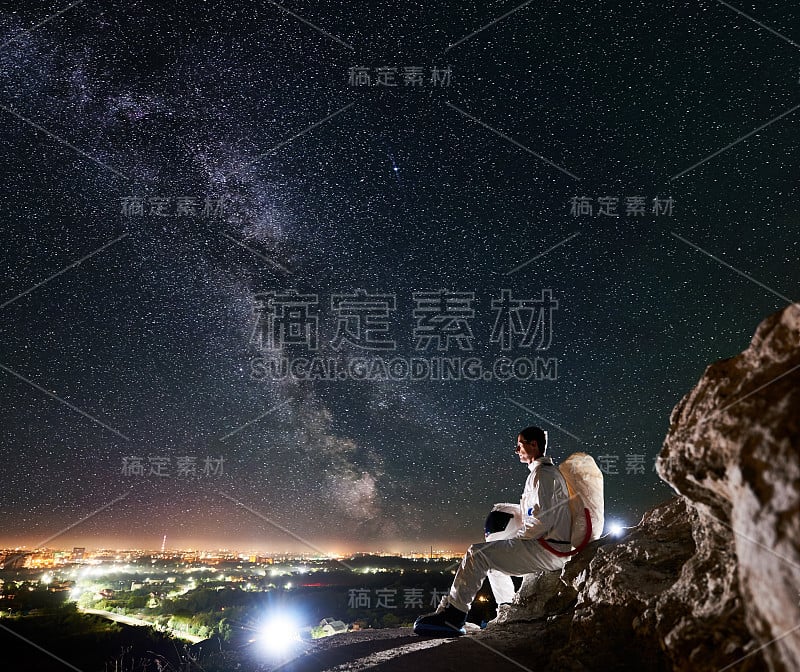 宇航员坐在布满星星的美丽夜空下的岩石山上。