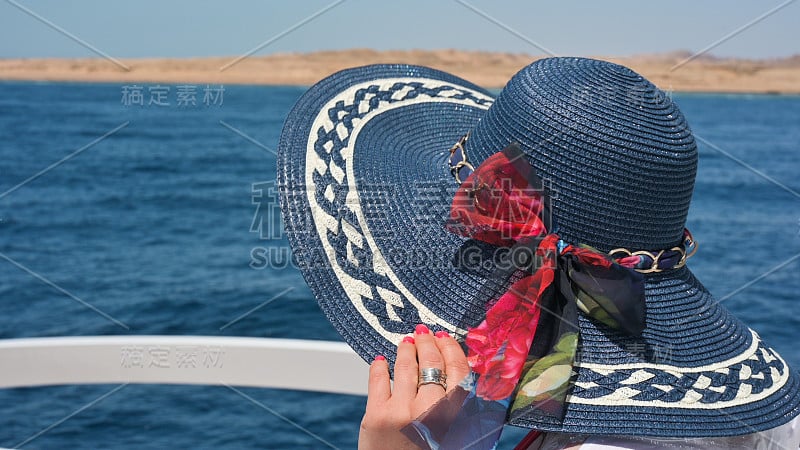 一个戴着宽边帽的女人坐在豪华快艇的甲板上，以大海为背景。在海上的游艇上戴着宽边帽的女人。脸是不可见的