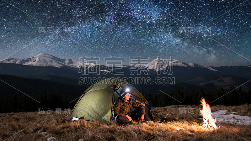 男游客晚上在山中露营休息。在满是星星和银河的美丽夜空下，一个带着头灯的男人坐在篝火和帐篷旁。图片宽高