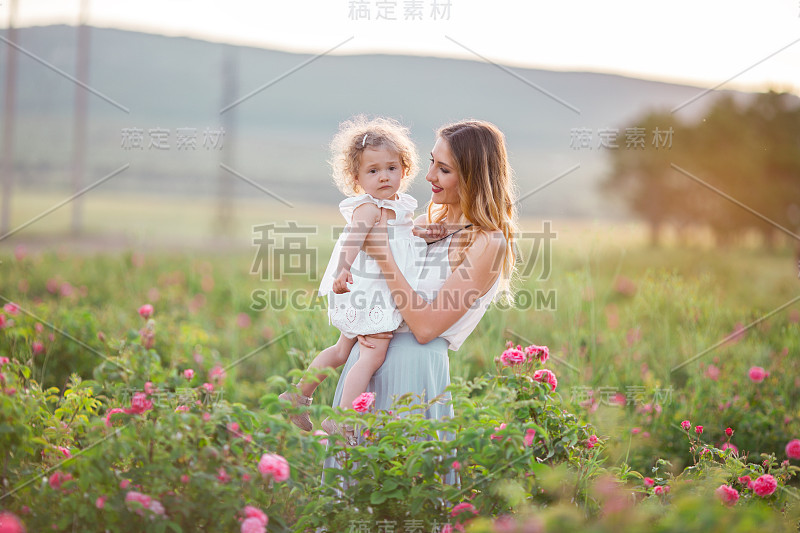 漂亮的孩子女孩和漂亮年轻的母亲穿着休闲的衣服，走在春天的花园与粉红色的花玫瑰