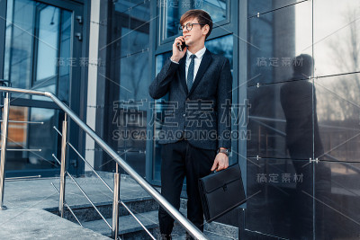 戴着眼镜的时尚商人在商务中心的玻璃大楼附近打电话