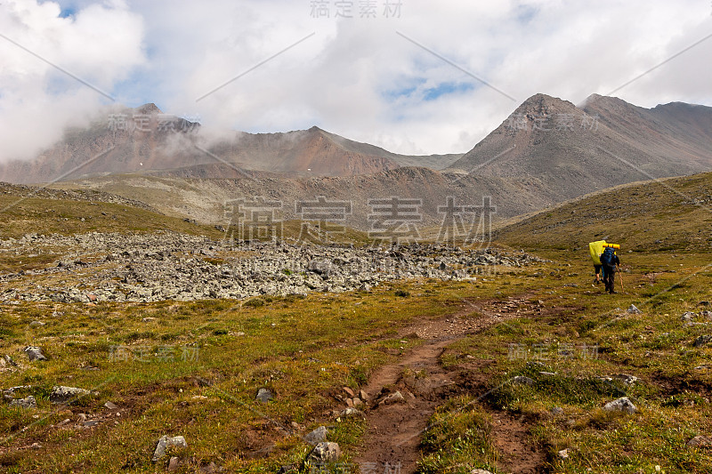 背着大背包的游客在山里徒步旅行。高山顶上的云朵。石头岩屑。山坡上有绿色的草。水平的。