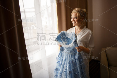穿着蓝色婚纱的新娘靠近窗户。婚礼的早晨。婚姻的概念