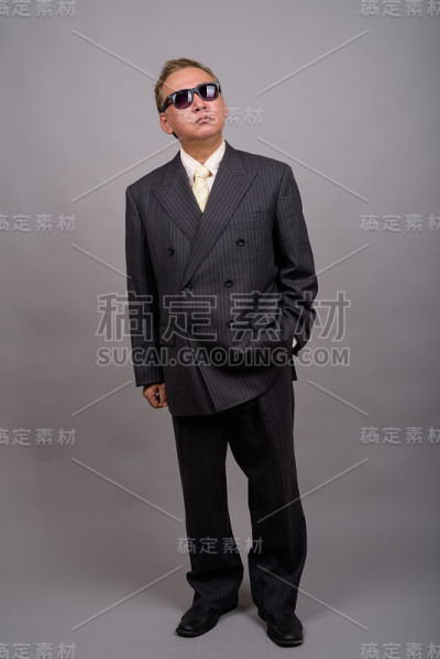 在灰色的背景下，成熟的亚洲商人戴着太阳镜看起来很酷