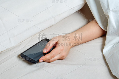 年轻女子躺在羽绒被下，拿着智能手机