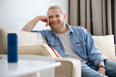 微笑的男人放松在沙发上看相机使用便携式无线扬声器在桌子上