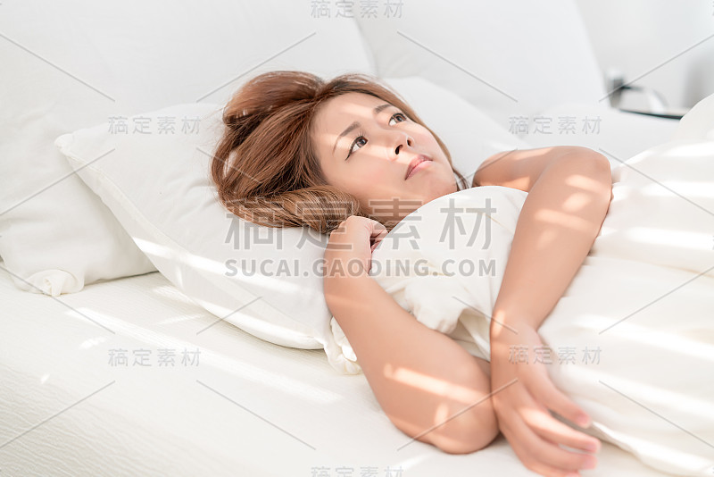 美丽的亚洲女人肖像与迷人的微笑享受新鲜柔软的床上用品亚麻布床垫在白色的卧室现代公寓。可爱的亚洲女孩睡