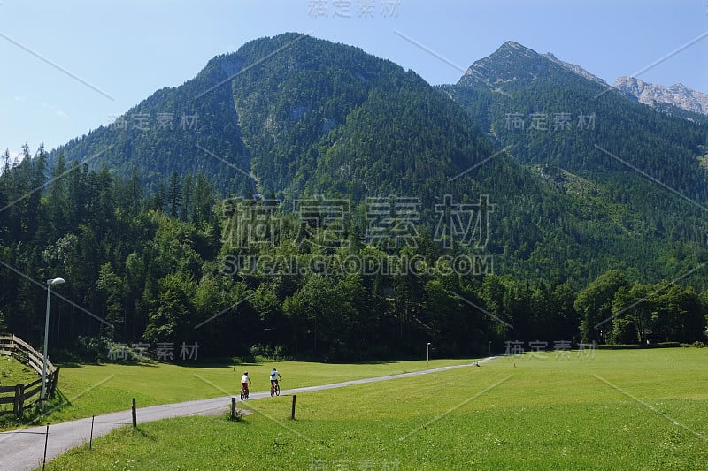 在阳光明媚的夏日里，两个人在草地之间的乡间小路上骑行的全景，背景是雄伟的群山