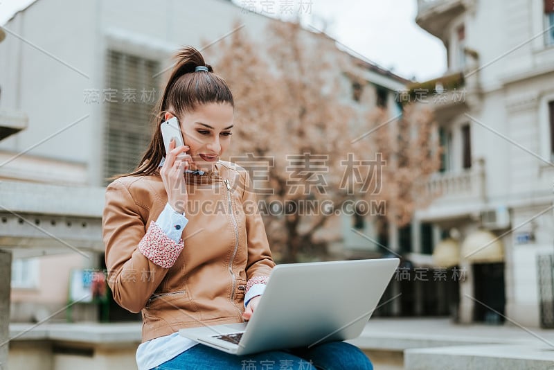 年轻漂亮的女人在户外使用她的笔记本电脑和手机。