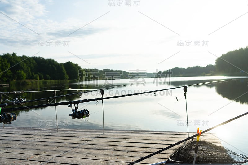 夏天的早晨，在美丽的蓝色湖泊上用鲤鱼竿和鱼荚钓鱼。在木制平台上钓鱼。