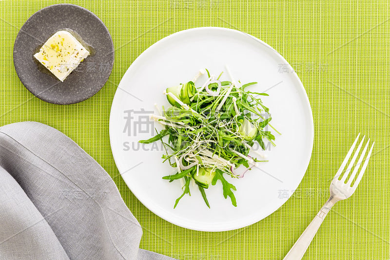 芝麻菜沙拉，配上豆芽、金针菇和黄瓜，配上羊乳酪，一张灰色的餐巾，银色的叉子搭在绿色的布料上。平的。素