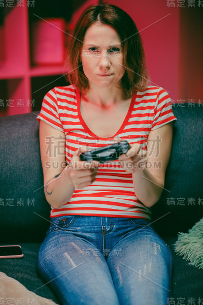 女孩晚上在客厅玩电子游戏。女玩家坐在沙发上，用无线控制器玩着视频游戏