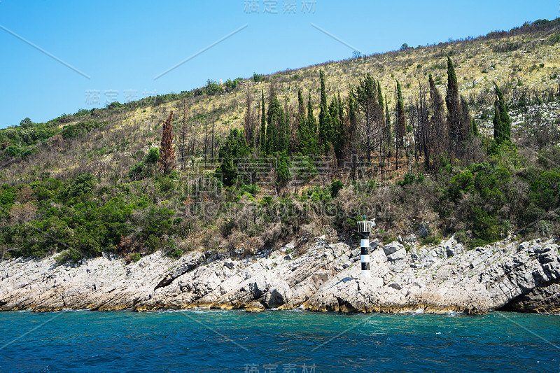 岩石海岸与一个小的黑白灯塔和森林与无云的天空在一个阳光明媚的夏日。科托尔湾。黑山。