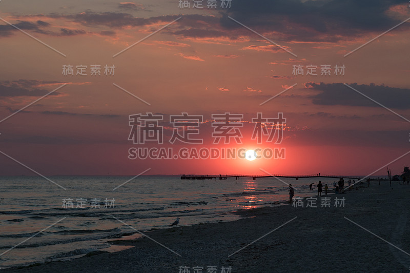 海滩上红色的夕阳。人们在海滩上剪影和海鸥。天际线上的码头。大红色的太阳