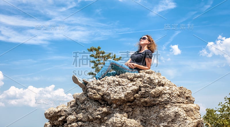 夏天在山顶上放松的女人。快乐美丽的人在悬崖上晒太阳。美丽的成年女孩休息后徒步在大自然的蓝天背景。