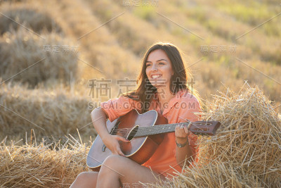 快乐快乐的女孩在日落时分的干草堆里弹吉他