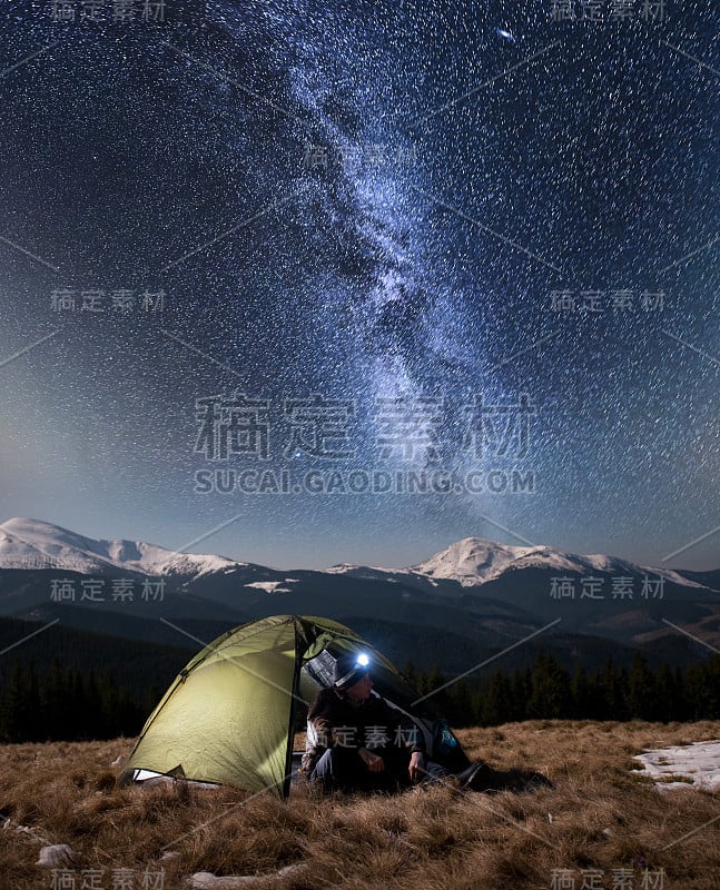 男徒步旅行者晚上在山里的露营中休息。在满是星星和银河的美丽夜空下，一名男子坐在旅游帐篷附近，欣赏夜景