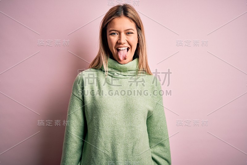 年轻美丽的金发女人穿着冬天的羊毛毛衣在粉红色孤立的背景伸出舌头快乐与有趣的表情。情感的概念。