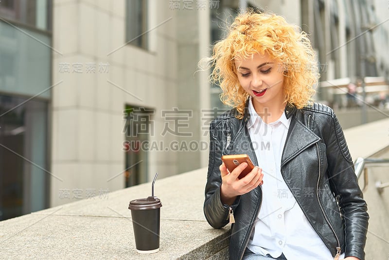 金发卷卷的年轻女子拿着纸玻璃杯和手机，背景是城市风景