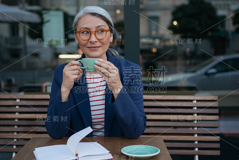 成熟的商业女性喝着咖啡，坐在咖啡馆里。沉思的亚洲女性的肖像戴着时尚的眼镜，拿着一杯茶