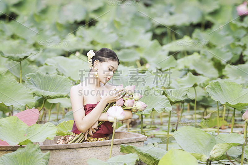 穿着泰国服装的妇女正在欣赏莲花。亚洲妇女坐在木船上采莲。美丽的女孩穿着泰国传统服饰，手拿鲜花。