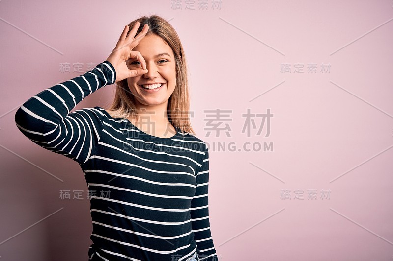 年轻美丽的金发女人穿着休闲条纹毛衣在粉红色孤立的背景做ok手势与手微笑，眼睛看着通过手指与幸福的脸。