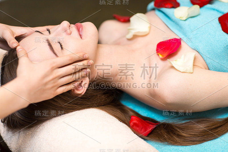 年轻美丽的女人接受头部面部按摩美容温泉与玫瑰花瓣