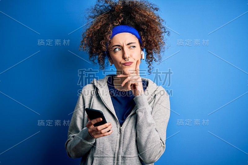 漂亮的女运动员带着卷发和穿孔穿运动服用智能手机严肃的面对思考问题，非常困惑的想法