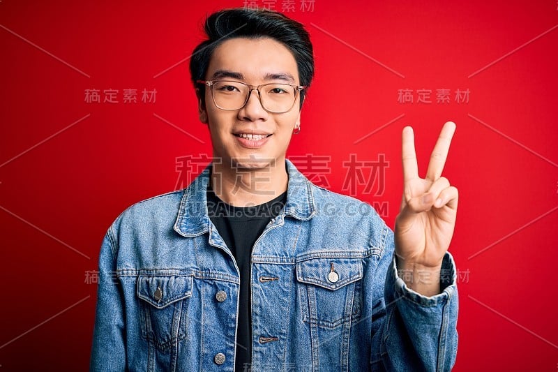 年轻帅气的中国男子穿着牛仔夹克，戴着眼镜，在红色背景下伸出食指，自信而快乐地微笑着。