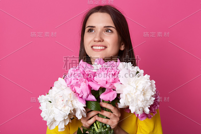 水平拍摄美丽的年轻女子与自然的化妆和深色长发拿着粉红色和玫瑰牡丹花，梦幻般的女性抬头，穿着黄色的服装