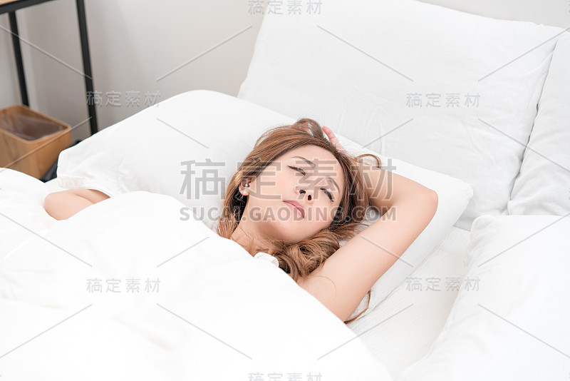 美丽的亚洲女人肖像与迷人的微笑享受新鲜柔软的床上用品亚麻布床垫在白色的卧室现代公寓。可爱的亚洲女孩睡