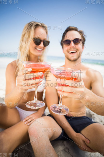 一对戴着太阳镜的年轻夫妇在海滩上展示鸡尾酒杯
