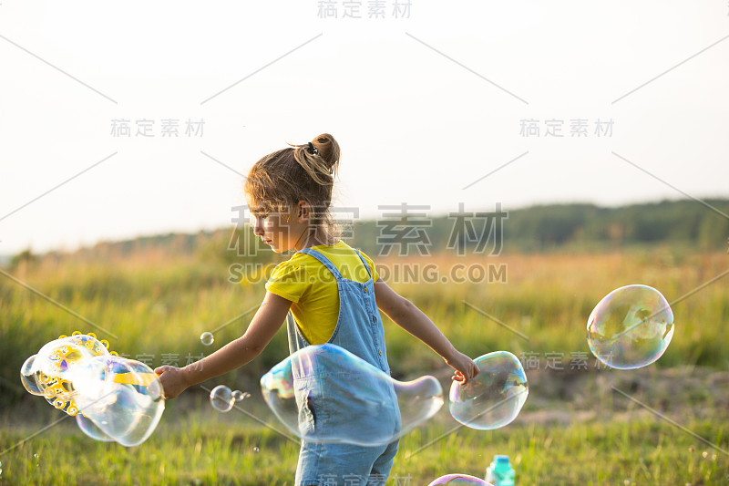 一个穿着牛仔连身裤的女孩在夏天的日落时分吹肥皂泡在田野里。国际儿童节，快乐儿童，户外活动。夏天的背景