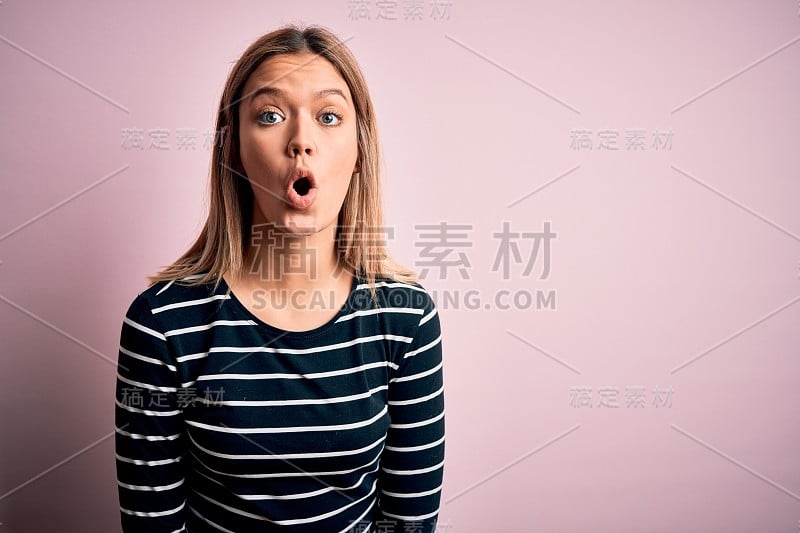 年轻美丽的金发女人穿着休闲条纹毛衣在粉红色孤立的背景害怕和震惊的表情，恐惧和兴奋的脸。