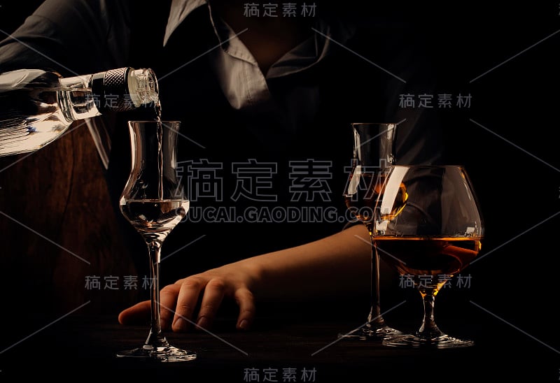 酒保将伏特加或龙舌兰酒倒入旧吧台上的小酒杯中。复古的木质背景在酒吧或酒吧，夜晚的心情。放置文本，色调