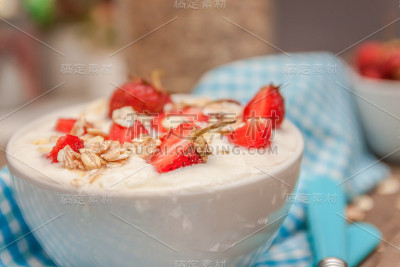 在一杯酸奶、燕麦格兰诺拉麦片和新鲜草莓中，在蓝色的汤匙毛巾上，散落的燕麦和草莓，在一个木制的背景上。