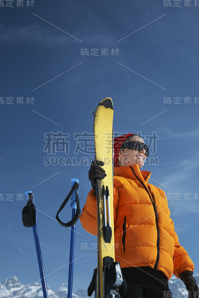 在蓝天下，穿着冬季夹克拿着滑雪板的女人