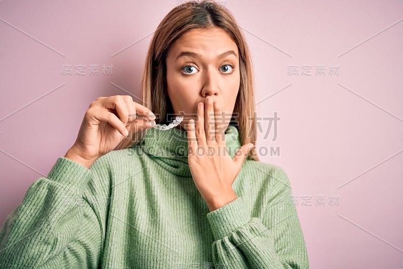 年轻美丽的女子抱着对准站在孤立的粉红色背景盖住嘴用手为错误而震惊，表情恐惧，在沉默中害怕，秘密的概念