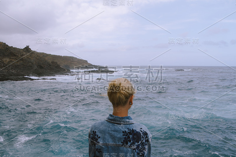 年轻女子坐在岩石上，看着汹涌的大海和破浪