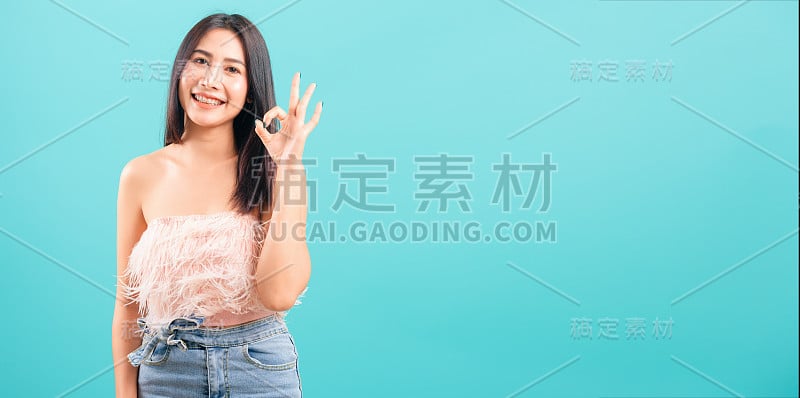 微笑的脸亚洲美丽的女人，她显示OK手势和看着相机在蓝色的背景