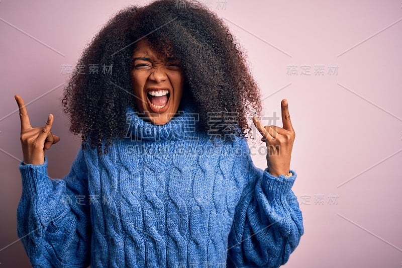 年轻美丽的非洲裔美国女人与非洲发式穿着冬季毛衣在粉红色的背景大喊与疯狂的表情做摇滚符号与手向上。音乐