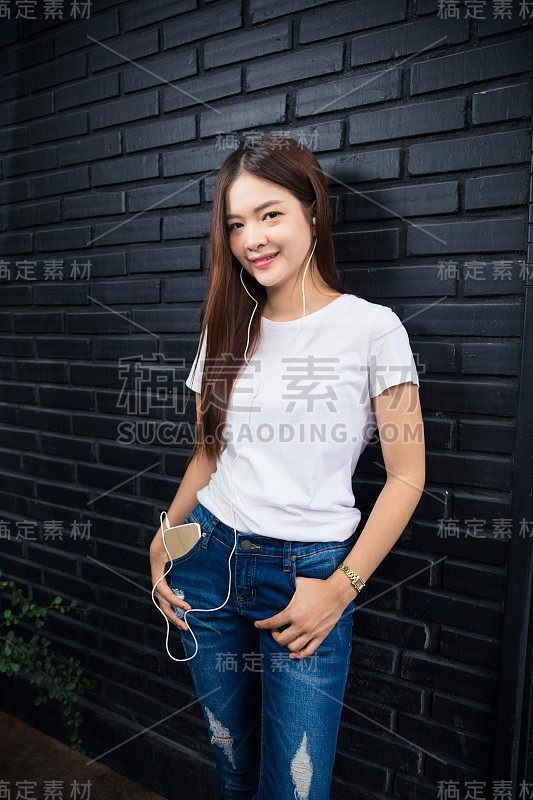 穿着白色t恤和蓝色牛仔裤的年轻快乐的女人靠在深色砖墙上，一边听着手机上的音乐，这是年轻潮人一代使用科